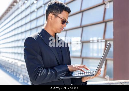 Junger Geschäftsmann, der an sonnigen Tagen mit einem Laptop eine Sonnenbrille trägt und am Geländer steht Stockfoto