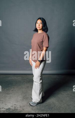 Selbstbewusste junge Frau, die vor grauer Kulisse im Studio steht Stockfoto