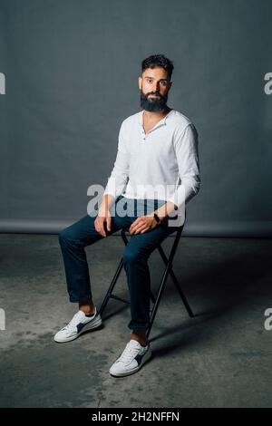 Selbstbewusster junger Mann mit Bart, der im Studio auf einem Stuhl sitzt Stockfoto