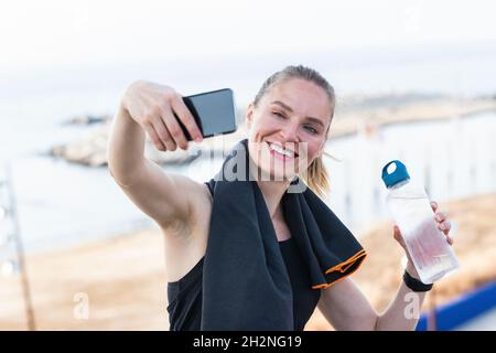Die Sportlerin nimmt Selfie über das Smartphone auf, während sie die Wasserflasche in der Hand hält Stockfoto