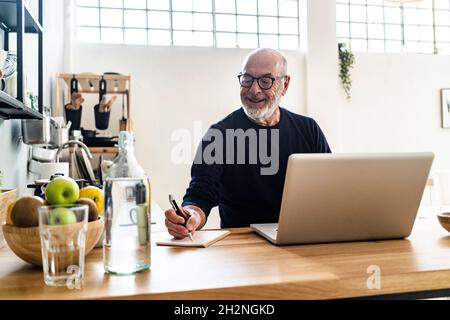 Älterer Mann, der einen Laptop benutzt, während er an der Küchentisch sitzt Stockfoto