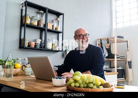 Glücklicher älterer Mann, der an der Küchentheke einen Laptop benutzt Stockfoto