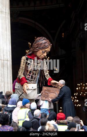 London, Großbritannien. Oktober 2021. Der Pastor von der St. Paul Cathedral erhält das Zeichen der Wertschätzung vom Little Amal.Little Amal, eine 3.5 Meter hohe Puppe, die als 9 Jahre altes syrisches Flüchtlingsmädchen dargestellt wird und alle Flüchtlingskinder vertritt, kam im Zentrum von London an der Großen Tür der St. Paul Cathedral an. Seit Juli dieses Jahres ist sie auf Reisen und verlässt die syrisch-türkische Grenze auf der Suche nach ihrer Mutter. Sie wird 8000 km laufen und Anfang November als ihr endgültiges Ziel in Manchester ankommen, wo sie auch den Standort der kurzfristigen Halteeinrichtung für Flüchtlinge in der ist Stockfoto