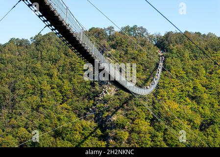 Eine hängende Holzbrücke mit Stahlseilen von der Seite aus gesehen gegen einen blauen Himmel, sichtbare Touristen auf der Brücke. Stockfoto