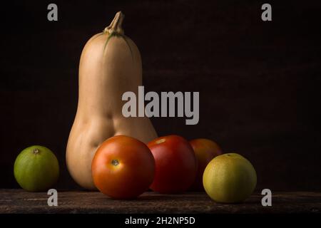 Verschiedene Bio-Gemüse auf einer dunklen, strukturierten Holzoberfläche, Seitenansicht von Butternusskürbis, Tomaten und Limette, rustikales Konzept mit Kopierraum Stockfoto