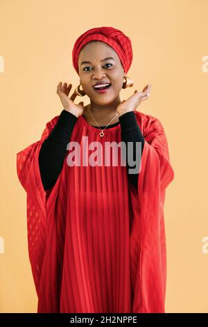 Schöne emotional lächelnde Frau mit hellem Make-up vor gelbem Hintergrund. Studioaufnahme eines weiblichen afrikanischen Models in ethnisch rotem Kleid und Schal, der die Kamera ansah und lächelte. Stockfoto
