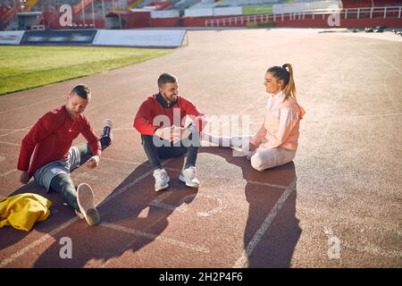 Junger erwachsener kaukasischer bärtiger Kerl, der zusammen mit einem lächelnden Paar auf der Sportlerstrecke sitzt, zu dem er spricht. Persönliches Trainerkonzept Stockfoto