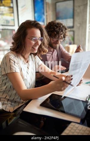 kaukasische Frau, die Papier hält, liest, lächelt und neben einem männlichen Kollegen im Büro sitzt. Stockfoto