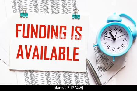Der Text ist auf einem Notizblock, der auf den Finanzdokumenten liegt, neben einem Stift und einem Wecker geschrieben. Sponsern Sie Finanzierungsgeschäfte Stockfoto