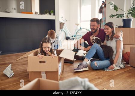 Familie im Wohnzimmer haben einen Spaß verbringen Zeit im neuen Zuhause.liebenswert verspielt lachende Mädchen sitzen an Karton, Junge rollt sie zusammen spielen. Stockfoto