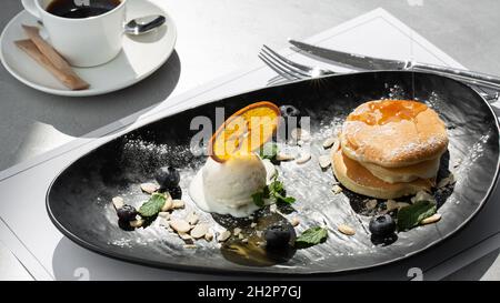 Amerikanische Pfannkuchen mit Eislöffel, Beeren und Ahornsirup auf schwarzem Teller isoliert mit Kaffee. Café-Menü Stockfoto