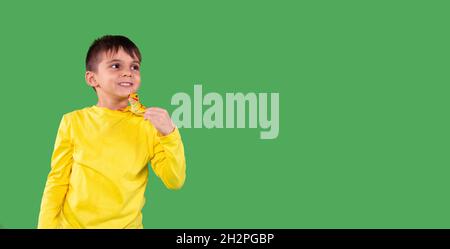 Foto eines lächelnden Jungen im Alter von Schulklassen mit gelbem Hoodie, der zur Seite schaut und das Dinosaurier-Spielzeug auf seiner Schulter vor dem grünen Hintergrund hält, leer Stockfoto