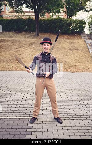 Full Body lustige männliche Darsteller in Kostüm lächelnd und jonglieren Messer, während auf dem Bürgersteig während der Show im Park stehen Stockfoto