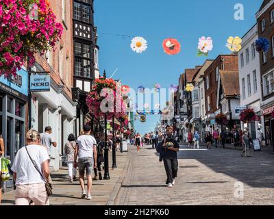 Geschäfte in der Guildford High Street mit Einkäufern, die teilweise Gesichtsmasken tragen. Blumenkörbe im Herbst und farbenfrohe Banner der Saison in klarem, blauem, warmem Sonnenschein Stockfoto