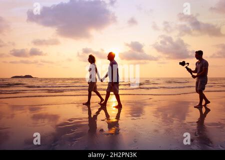 Filmemacher, der am Strand bei Sonnenuntergang ein Paar mit Kamerastabilisator filmt, und professioneller Videofilmer, der am Film arbeitet Stockfoto