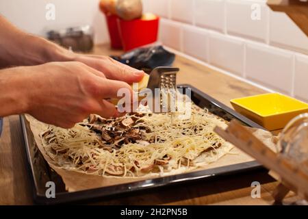 Nahaufnahme der Hände Reiben Käse auf der Pizza oder Flammekueche mit Pilzen, Kochen in der heimischen Küche Stockfoto