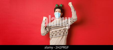 Konzept von Coronavirus, Winterurlaub und sozialer Distanzierung. Bild von aufgeregt und glücklich Mann in medizinische Maske und weihnachtskleidung, die Hände nach oben zu heben Stockfoto