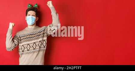 Konzept von Coronavirus, Winterurlaub und sozialer Distanzierung. Bild von aufgeregt und glücklich Mann in medizinische Maske und weihnachtskleidung, die Hände nach oben zu heben Stockfoto