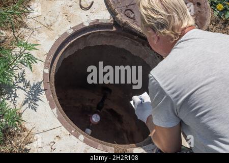 Ein Mann zeichnet die Messwerte des Wasserzählers auf und lehnt sich über den Brunnen. Prüfung und Abrechnung von Messgeräten. Stockfoto