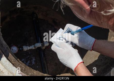 Ein Mann zeichnet die Messwerte des Wasserzählers auf und lehnt sich über den Brunnen. Prüfung und Abrechnung von Messgeräten. Stockfoto