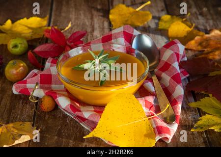 Kürbissuppe in einer Glasschüssel auf einem Holztisch, geschmückt mit bunten Herbstblättern und kleinen wilden Äpfeln Stockfoto