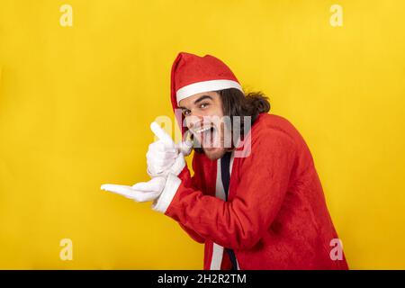 Ein lächelnder junger Mann, der als Weihnachtsmann gekleidet ist, zeigt mit der Hand auf Ihr Objekt und hebt den Daumen zur Zustimmung. Gelber Hintergrund und Kopierplatz für Stockfoto