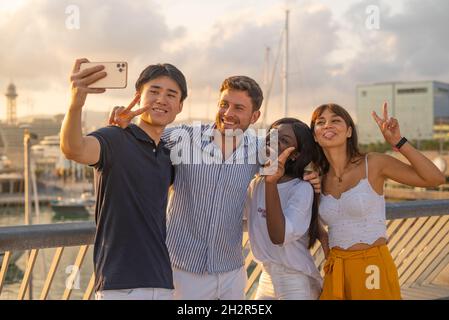 Unbeschwerte junge, vielfältige Freunde in lässigen Outfits, die lächeln und Friedensgesten machen, während sie am Sommerabend Selfie auf dem Smartphone während der gemeinsamen Freizeit am Ufer machen Stockfoto
