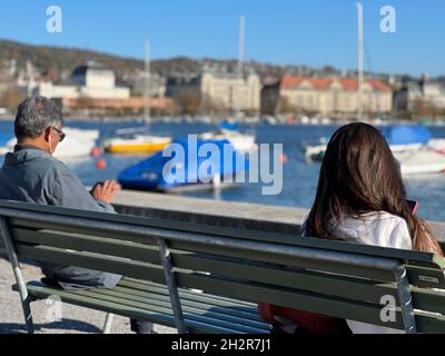 Frau und Mann sitzen auf einer Holzbank und halten sich sozial distanziert. Die sitzen auf gegenüberliegenden Enden der Bank und jeder von ihnen schaut in ihre Telefone. Stockfoto