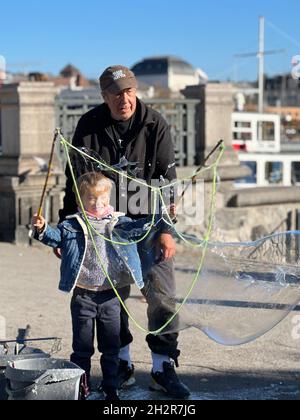 Der alte Mann hilft einem kleinen Jungen dabei, riesige Seifenblasen zu erzeugen. Der Junge hält zwei Stöcke, die mit einer Schnur verbunden sind. Es gibt mehrere riesige Blasen. Stockfoto