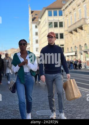 Interrassisches Paar beim Spazierengehen auf der Straße und beim gemeinsamen Genießen im Stadtzentrum von Zürich in der Schweiz. Der Mann hat eine Papiertasche. Stockfoto