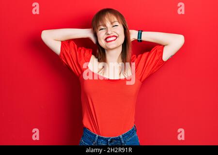 Rotschopf junge Frau trägt lässiges rotes T-Shirt zum Entspannen und Stretching, Arme und Hände hinter Kopf und Hals lächeln glücklich Stockfoto