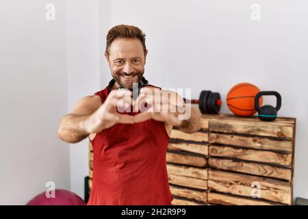 Mann mittleren Alters trägt Sportkleidung und Kopfhörer im Fitnessstudio lächelt verliebt und zeigt Herz-Symbol und Form mit den Händen. Romantisches Konzept. Stockfoto
