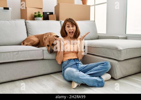 Junge kaukasische Mädchen in legerer Kleidung sitzt auf dem Boden mit Hund zu Hause feiern Sieg mit glücklichen Lächeln und Sieger Ausdruck mit Rais Stockfoto