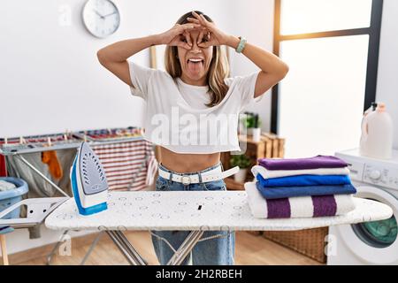 Junge hispanische Frau bügelt Kleidung in der Waschküche tut ok Geste wie Fernglas aufragende Zunge aus, Augen durch die Finger schauen. Crazy Express Stockfoto
