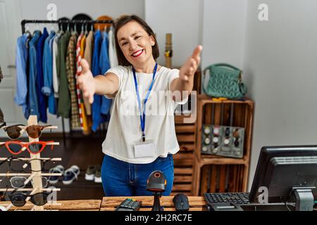 Schöne hispanische Frau im mittleren Alter, die als Managerin in einer Einzelhandelsboutique arbeitet und die Kamera lächelt, um sich mit offenen Armen zu umarmen. Fröhlicher Ausdruck em Stockfoto