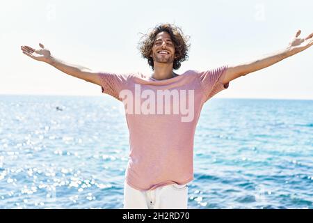 Junger hispanischer Mann atmet beim Stehen mit offenen Armen am Strand. Stockfoto