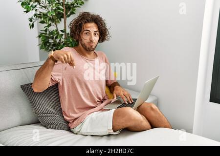Ein junger hispanic-Mann, der zu Hause auf dem Sofa sitzt und mit einem Laptop nach unten zeigt, traurig und aufgeregt aussieht und mit Fingern die Richtung angibt, unglücklich und depres Stockfoto