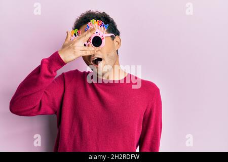 Hübscher hispanischer Mann, der eine lustige Geburtstagsbrille trägt, die mit der Hand ins Gesicht und in die Augen blickt und Angst durch die Finger schaut Stockfoto