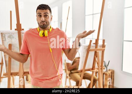 Junger hispanischer Mann im Kunststudio ahnungslos und verwirrt Ausdruck mit Armen und Händen angehoben. Zweifel Konzept. Stockfoto