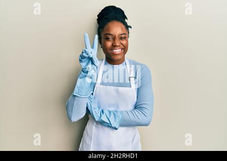 afroamerikanische Frau mit geflochtenen Haaren, die eine Reinigungsschürze trägt, und lächelnden Handschuhen mit einem glücklichen Gesicht, das die Kamera beim Siegeszeichen anwinkelt. Nummer zwei Stockfoto