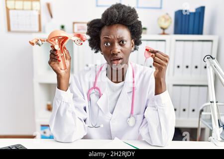 Afrikanische Ärztin, die anatomisches weibliches Genitalorgan und Menstruationsbecher im Schockgesicht hält, skeptisch und sarkastisch aussieht, überrascht mit offenem mou Stockfoto