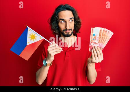 Junger hispanischer Mann mit philippinischen Flaggen und Pesos-Banknoten, der lächelnd zur Seite schaut und denkend wegstarrt. Stockfoto