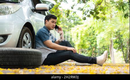 Asiatischer Mann, der neben dem Auto sitzt und nach einer Panne auf der Straße mit dem Mobiltelefon um Hilfe telefoniert. Konzept des Motorproblems oder der Accid des Fahrzeugs Stockfoto