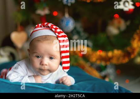 Porträt eines neugeborenen Kindes in einer gestreiften Mütze vor dem Hintergrund eines Weihnachtsbaums. Erstaunt neugeborenes Baby mit schönen blauen Augen Blick auf c Stockfoto