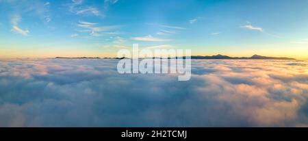 Luftaufnahme des leuchtend gelben Sonnenaufgangs über weißen dichten Wolken und fernen Bergen am Horizont. Stockfoto