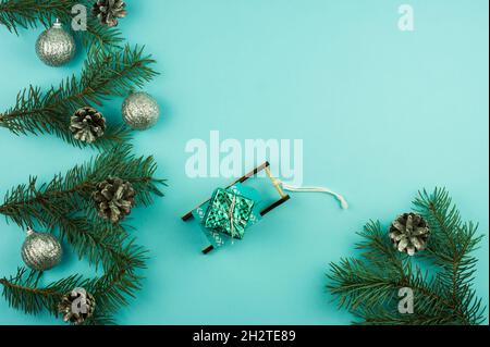 Neujahrskomposition mit dekorierten Zweigen aus Fichtensilberkugeln und Zapfen. Dekorativer Schlitten mit einem Geschenk auf blauem Hintergrund. Kopierraum Stockfoto