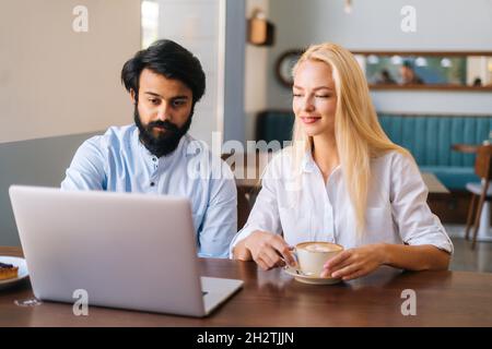Vorderansicht von zwei Geschäftsleuten eines bärtigen indischen Mannes und einer attraktiven kaukasischen blonden Frau, die mit einem Laptop im Café arbeitet und Kaffee trinkt. Stockfoto