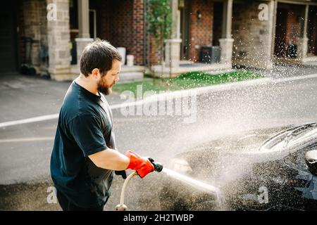 Cacuasian junger Mann, der Auto mit dem Wasserstrahl vor dem Haus zu Hause wäscht. Stockfoto