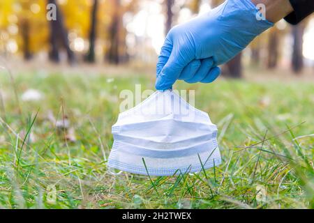 Nahaufnahme einer Hand, die einen Einweghandschuh trägt und eine weggeworfene Gesichtsmaske vom Boden im örtlichen Park aufnimmt. Coronavirus-Kunststoffabfälle verschmutzen Stockfoto