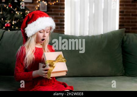 Kleine kleine schockierte Mädchen in roten Kleid hält Weihnachten oder Neujahr Geschenk-Box. Kaukasisches Kind überrascht und sagen wow emotional, verlassen, weil Stockfoto